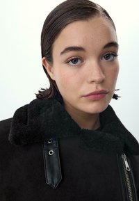 Czarna kurtka z futrzanym kołnierzem, skórzanymi akcentami i srebrnym zamkiem. Twarz modela jest częściowo widoczna, z gładką skórą i piegami na policzkach.