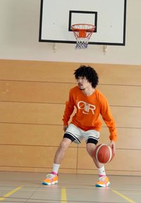 Chemise à manches longues orange avec un logo circulaire, shorts blancs et baskets multicolores. Jouant au basketball sur le sol d'un gymnase sous un panier.