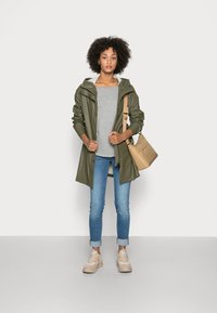 Olijfgroene waterdichte jas met een capuchon, grijs shirt, lichtblauwe jeans en beige sneakers; met een beige schoudertas.