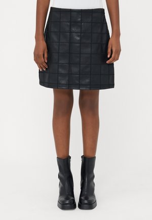 Mini-jupe en cuir noir avec motif carré cousu portée avec des bottines en cuir noir sur une personne debout.