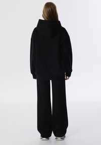 Sudadera negra de gran tamaño con capucha y puños acanalados, combinada con pantalones negros de pierna ancha. Textura de tela suave.