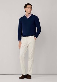 Granatowy sweter z dekoltem w serek nałożony na różowo-białą koszulę w paski z kołnierzykiem, do tego kremowe spodnie i brązowe buty. Prostota w klasycznym stylu.