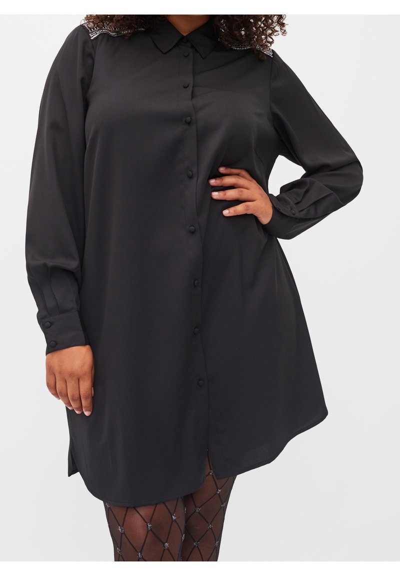 Robe noire à manches longues boutonnée avec col et épaules ornées, portée avec des collants transparents à motifs, main du modèle sur la hanche.