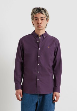 Giovane con capelli corti striati di chiaro, che indossa una camicia viola con bottoni e jeans blu, in piedi davanti a uno sfondo chiaro e uniforme.
