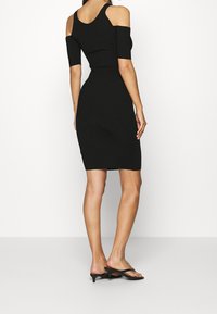 Femme portant une robe noire en maille ajustée avec des découpes aux épaules, se tenant de dos, associée à des sandales noires à talons bas.