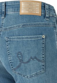 Jeans aus Denim mit einer hellen Waschung, versehen mit einem braunen Etikett mit wiederholtem Text auf der Rückseite. Beachten Sie die blauen Nähte an der Gesäßtasche.