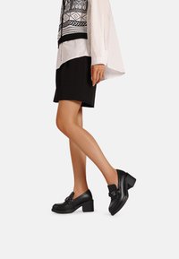 Scarpe nere in pelle con tacco, punta arrotondata, dettaglio decorativo con fibbia e tacco spesso. Il materiale ha una consistenza liscia e lucida.