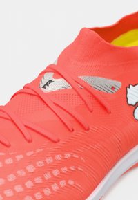 Jaskrawy koralowy but sportowy z teksturowaną cholewką, ozdobiony srebrnymi akcentami, sznurowadłami oraz charakterystycznym wzorem na przedniej części.