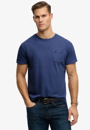 Uomo con capelli corti che indossa una maglietta blu girocollo con taschino sul petto, jeans scuri, cintura nera e orologio da polso, in piedi con una mano in tasca.