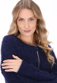 Młoda kobieta z długimi falującymi blond włosami, ubrana w granatowy miękki sweter, z założonymi rękami, pewnie patrzy w obiektyw na tle białego tła.