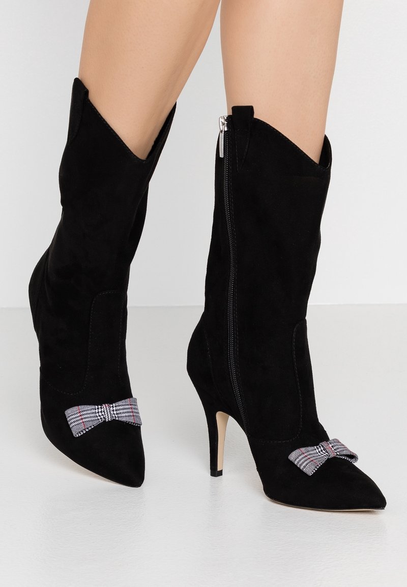 Bottines en suede noir avec des pointes et des talons hauts. Elles présentent une fermeture éclair sur le côté et un accent nœud tartan sur le devant.