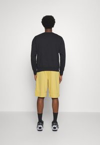 Sudadera negra con puños y dobladillo acanalados, combinada con pantalones cortos amarillos. Observada desde atrás, mostrando un ajuste relajado y un estilo casual.
