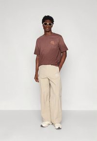 Brown cotton t-shirt with a small graphic, paired with beige wide-leg trousers. The model wears light sunglasses and neutral sneakers.