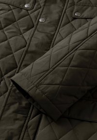 Quiltana jakna maslinaste boje s glatkom teksturom, metalnim dugmadi na pritisak i dijagonalnim šavovima na tkanini.