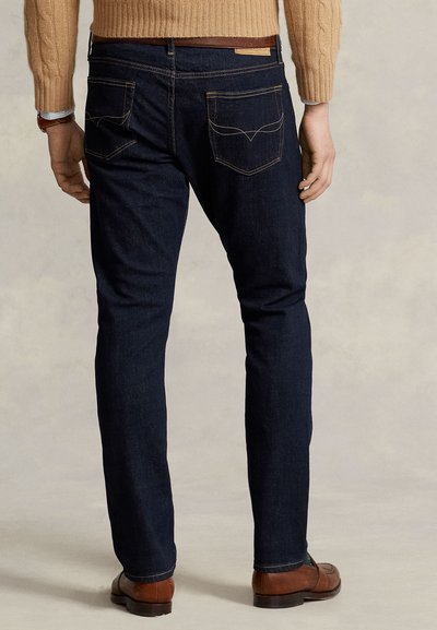 Hombre con vaqueros azul oscuro, suéter marrón claro y zapatos de cuero marrones, de pie de espaldas sobre un fondo liso.