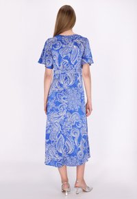 Femme aux longs cheveux raides portant une robe midi bleue avec des motifs paisley blancs et des sandales à talons argentées, se tenant de dos.