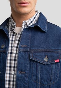 Denimska jakna temno modre barve, z dvema žepoma s poklopcem in s srebrnim okovjem. Nosena je čez črno-belo karirano srajco.