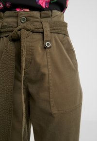 Olivgrön, hög midja byxor med vävd bälte, sidofickor och knapplies. Tillverkade av kraftigt bomullsmaterial med en slät textur.