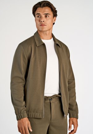 Uomo che indossa una giacca verde oliva con zip e pantaloni abbinati, con una t-shirt bianca, in piedi davanti a uno sfondo chiaro e uniforme, guarda verso sinistra.