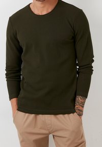 Maglietta a maniche lunghe verde oliva a coste con scollatura rotonda. Il tessuto è testurizzato. La persona indossa pantaloni beige, con tatuaggi visibili sulle braccia.