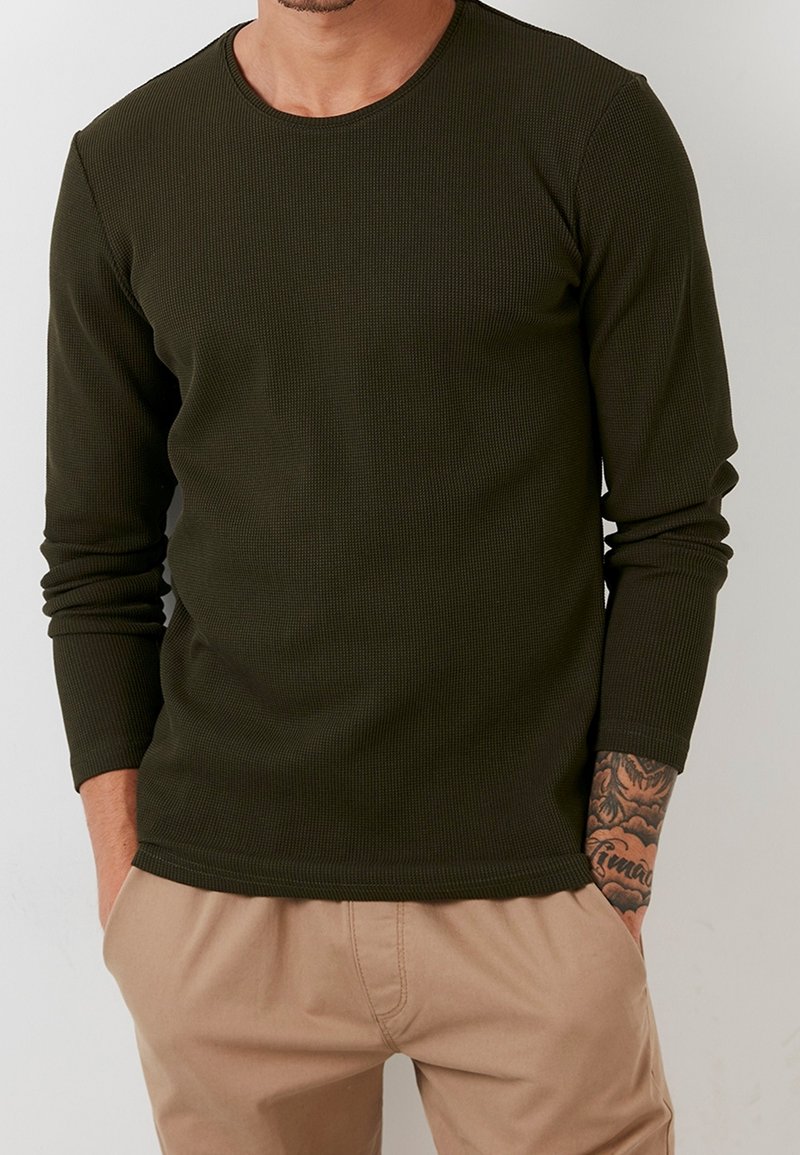 Maglietta a maniche lunghe verde oliva a coste con scollatura rotonda. Il tessuto è testurizzato. La persona indossa pantaloni beige, con tatuaggi visibili sulle braccia.