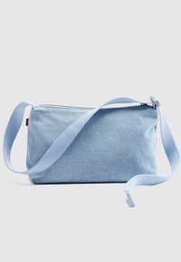 Τσάντα χιαστί από ανοιχτό μπλε τζην με ρυθμιζόμενο λουράκι και επάνω φερμουάρ, τοποθετημένη πάνω σε απλό ανοιχτό γκρι φόντο.