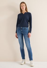 Haut à manches longues bleu marine avec col en V et patte de boutonnage à l'avant, associé à un jean droit bleu clair et des baskets blanches à lacets.