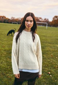 Krém színű, kötött mintás pulóver laza szabással és bordázott szegéllyel, fehér ingre húzva. Fekete csipkés szegélyű szoknya látható alul. Zöld fű háttér.