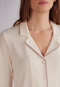 Crème-kleurige pyjama top met een geknipte kraag, donkerbordeaux piping en twee knopen aan de voorkant. Zachte stof met een gladde textuur.