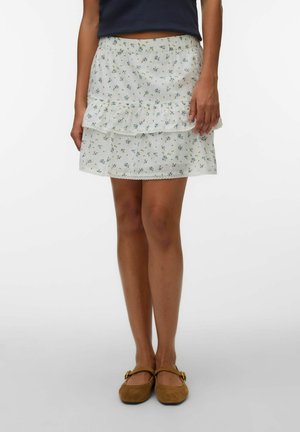 Femme portant une mini-jupe blanche à fleurs avec des volants, des ballerines marron et un top court bleu marine, debout devant un fond uni.