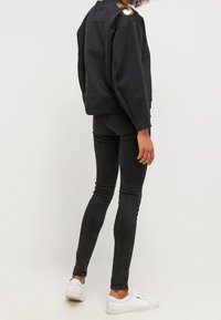 Sweat-shirt noir avec un col rond et un accent doré sur l'épaule, associé à un jean skinny noir et des baskets blanches.