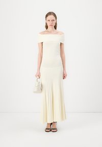 Robe crème décolletée, avec texture côtelée, jupe plissée évasée, sandales noires à lanières et un petit sac à main blanc.