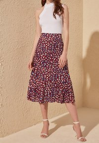 Nainen, jolla on yllään hihaton valkoinen halterneck-toppi, punakuvioinen midi-hame ja valkoiset nilkkaremmitossut, taustalla teksturoitu vaalea seinä.