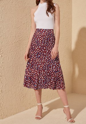 Woman wearing sleeveless white halter top, red patterned midi skirt, and white ankle-strap heels against textured beige wall.