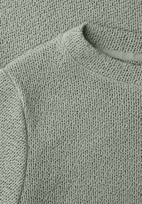 Jersey de punto gris claro con patrón texturizado tipo gofre, primer plano que muestra el escote, el hombro y parte de la manga.