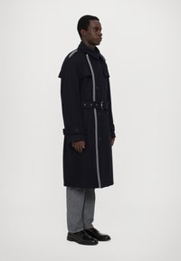 Trench coat blu scuro con accenti grigi, design doppiopetto, colletto ampio, vita con cintura e polsini abbottonati; abbinato a jeans grigi e scarpe nere.