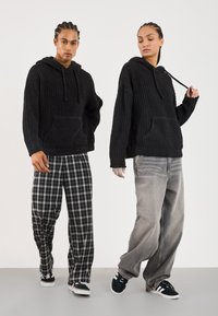 Zwarte gebreide hoodies hebben voorzakken en geribbelde manchetten, gecombineerd met zwart-witte geruite broeken en lichtgrijze jeans. Sneakers maken de outfits compleet.
