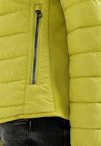 Gros plan d'une veste matelassée vert citron avec une poche à fermeture éclair noire verticale sur la poitrine, portée sur un jean denim foncé.