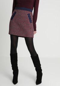 Neulottu minihame, jossa on punavalkoinen ja laivastonsininen ruutukuosi, laivastonsininen vyötärönauha ja kaksi laivastonsinistä taskua. Yhdistetty mustien polven korkeiden saappaiden kanssa.