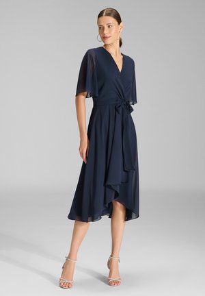Femme portant une robe midi bleu marine avec manches flottantes, col en V, ceinture à la taille et ourlet asymétrique, associée à des talons argentés à brides.
