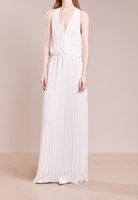 Kobieta ubrana w białą, bezrękawnikową suknię maxi z głębokim dekoltem w kształcie V i plisowanym dołem, stojąca na tle jednolitego, jasnobeżowego tła.