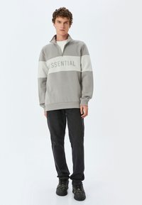 Grå zip-up sweatshirt med vit rand och texten "ESSENTIAL" över bröstet, kombinerad med svarta jeans och mörka skor.
