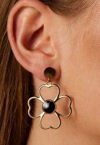 Blumenförmige Ohrringe in Goldfarbe mit einer schwarzen zentralen Perle, die eine kreisförmige Rückseite und eine zarte Umrandung aufweisen.