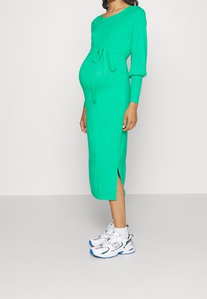 Vestido midi verde con mangas largas, abertura lateral y detalle de cinturón anudado, combinado con zapatillas blancas. Textura suave y diseño ajustado.