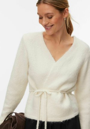 Femme portant un pull cache-cœur blanc doux avec une ceinture nouée, souriante et regardant vers le bas, tenant un sac marron dans une main.
