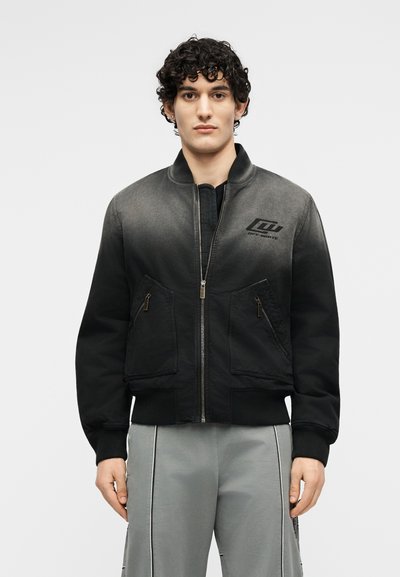 Νεαρός άνδρας με σγουρά μαλλιά φοράει μαύρο bomber μπουφάν με φερμουάρ και μπροστινές τσέπες, καθώς και γκρι παντελόνι με κάθετες ραφές.