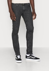 Jeans skinny de denim preto com uma textura suave, apresentando bolsos frontais, fecho zip e combinados com ténis preto e branco.