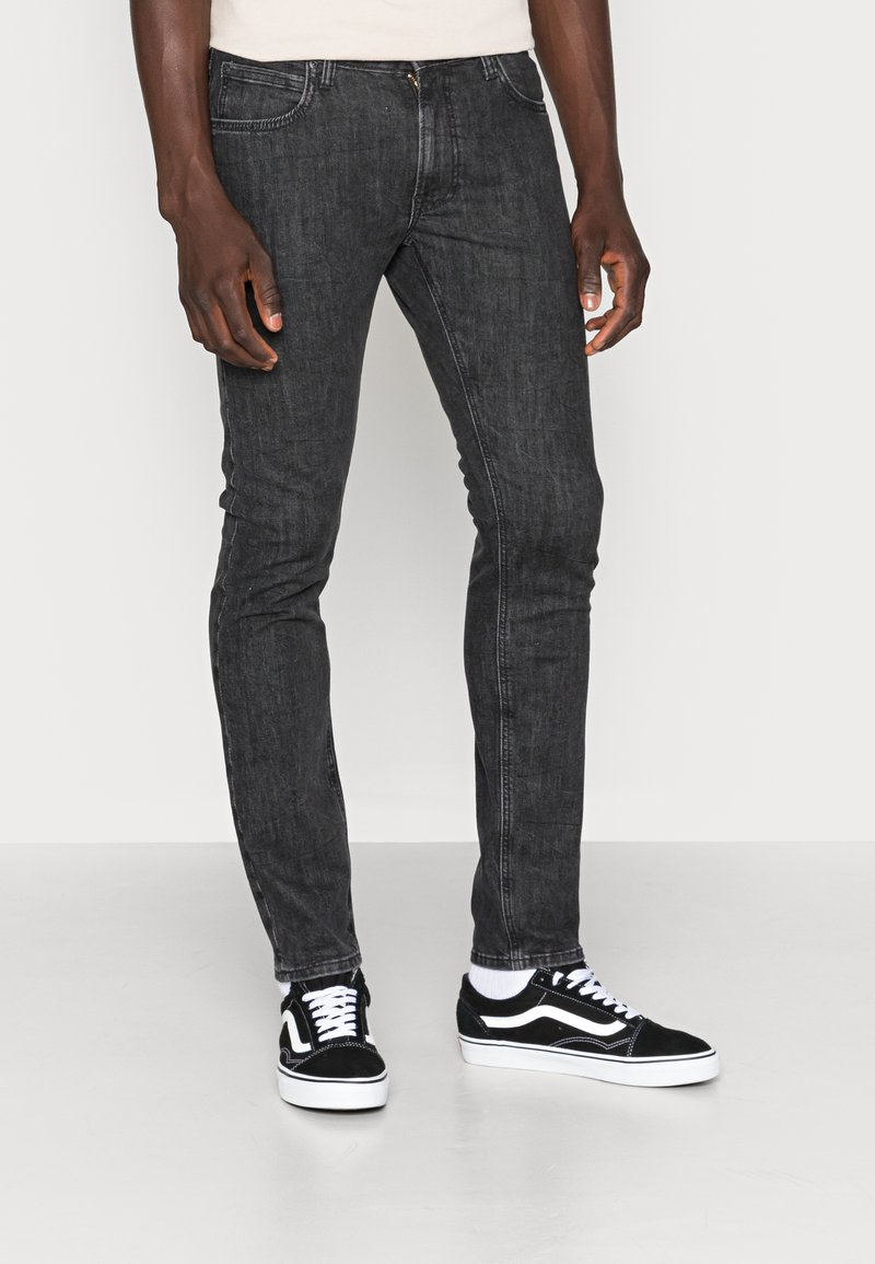 Jeans skinny de denim preto com uma textura suave, apresentando bolsos frontais, fecho zip e combinados com ténis preto e branco.