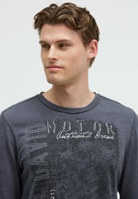 Graues Langarmshirt mit Rundhalsausschnitt, das auf der Vorderseite kontrastierende grafische Schriftzüge und Motordesign-Elemente aufweist. Glatte Textur.