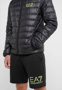 Giacca a vento nera imbottita con texture trapuntata e chiusura con zip. Presenta il logo verde EA7 sul petto a sinistra. Abbinata a pantaloncini neri con logo.